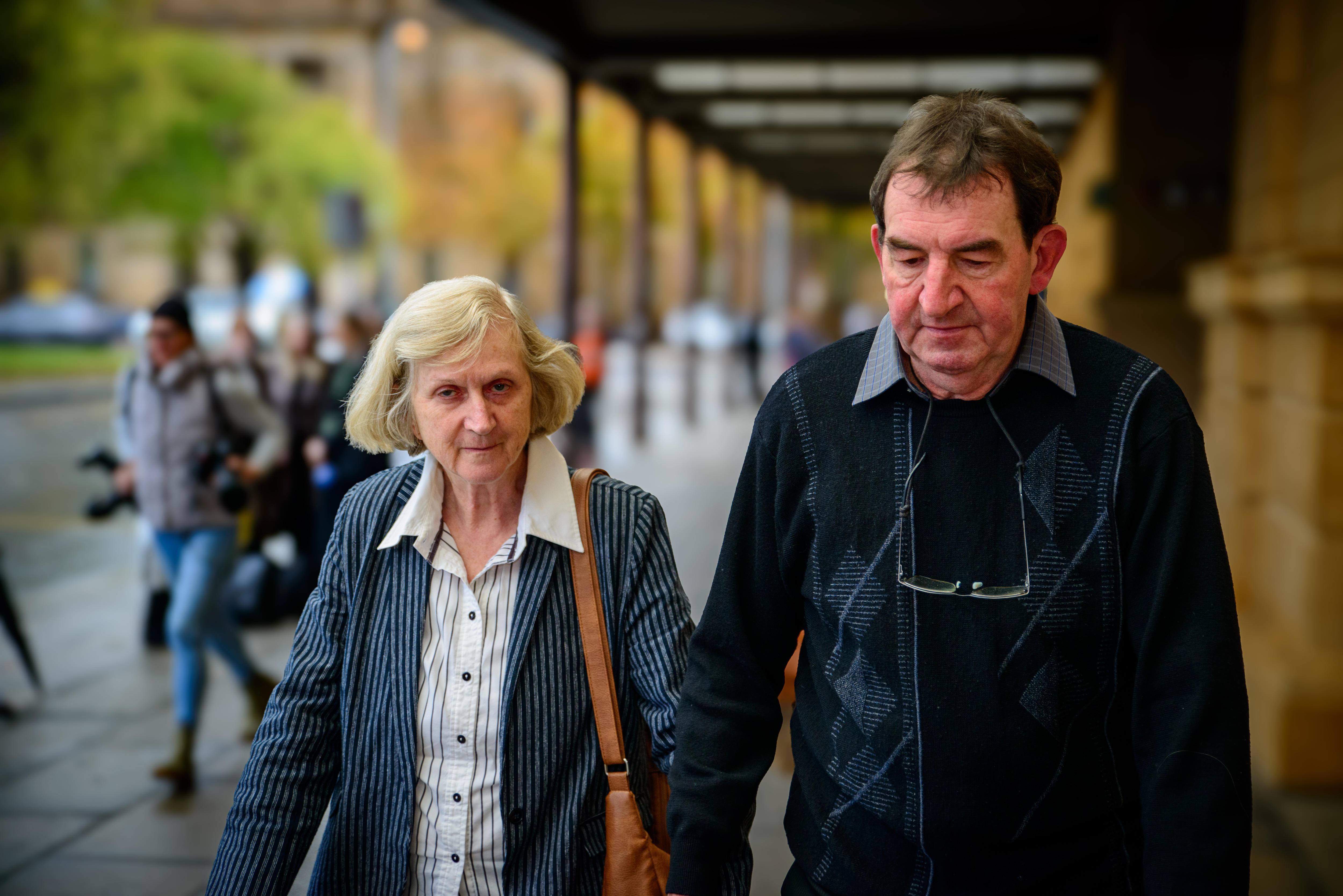 A man with glasses hanging around his neck is walking along a footpath holding hands with a woman.