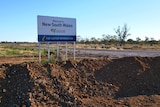 A sign that says welcome to New South Wales is surrounded by a mound of dirt on the road between both states. 
