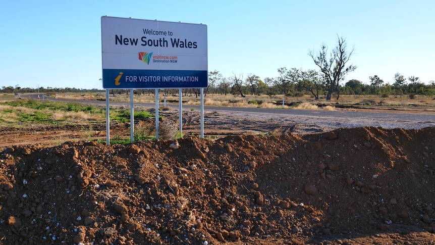A sign that says welcome to New South Wales is surrounded by a mound of dirt on the road between both states. 