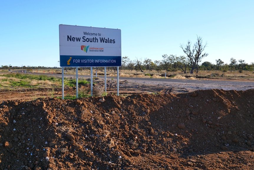 A sign that says welcome to New South Wales is surrounded by a mound of dirt on the road between both states. 