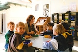 A group of young children sit around a table in a childcare centre with a female carer.