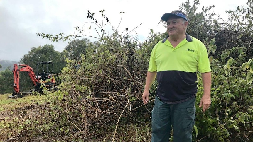 Gary Hudson standing in front of a pile of lantana and a machine that took it off the hill.