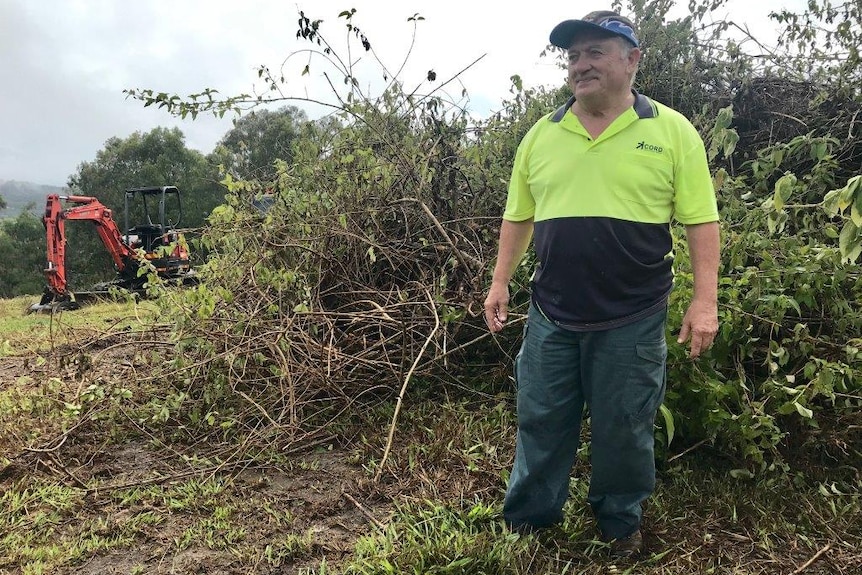 A man stands in front of a pile of lantana and a machine that took it off the hill.