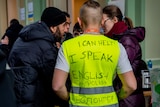 A man wearing a hi-vis vest with the message I SPEAK ENGLISH AND POLISH written on it speaks with a group