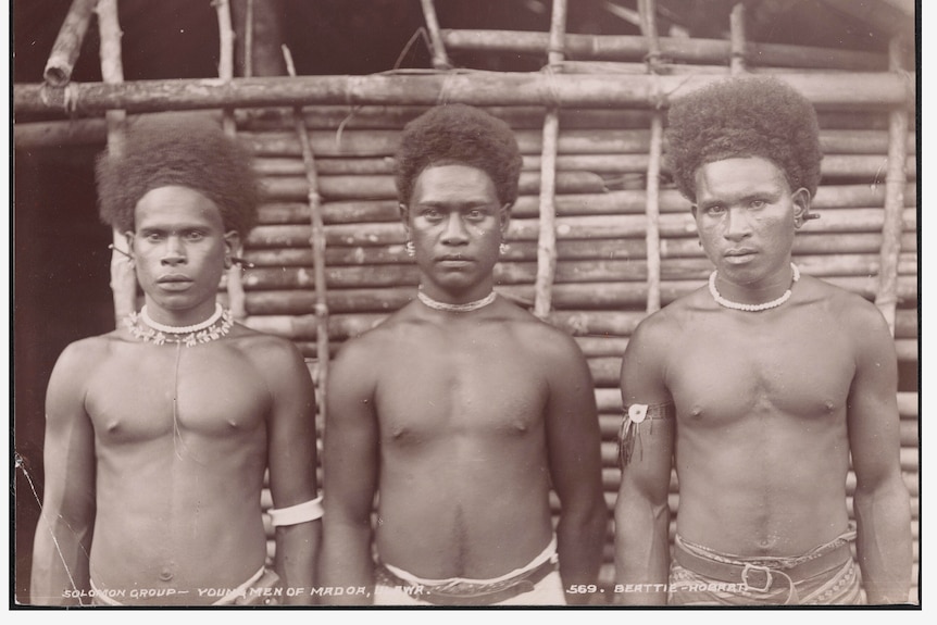 Three shirtless men from the Solomon Islands' Ulawa people stand outside a traditional building.
