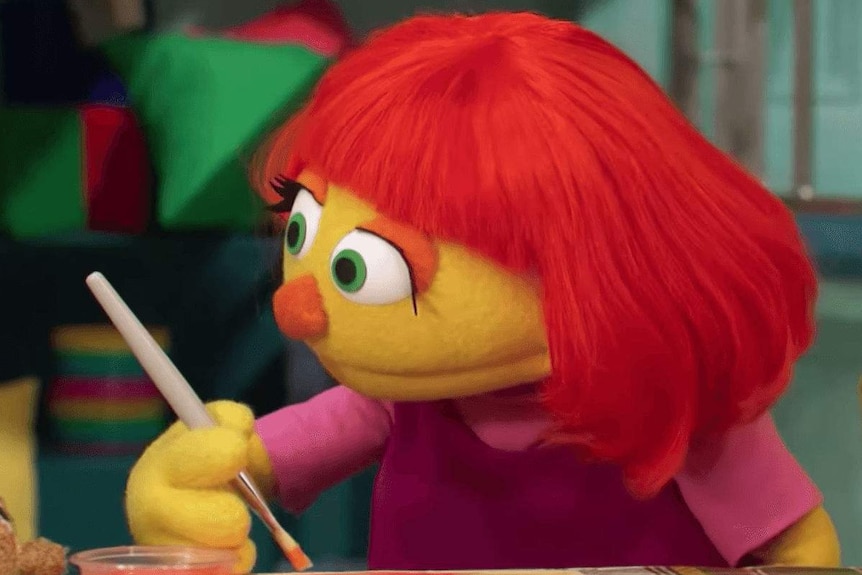 A female muppet with red hair and yellow skin sits at a table painting, she is wearing a pink T-shirt.