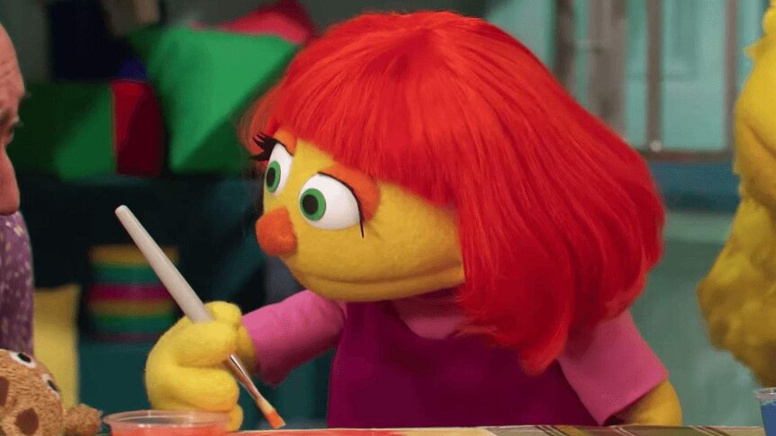 A female muppet with red hair and yellow skin sits at a table painting, she is wearing a pink T-shirt.