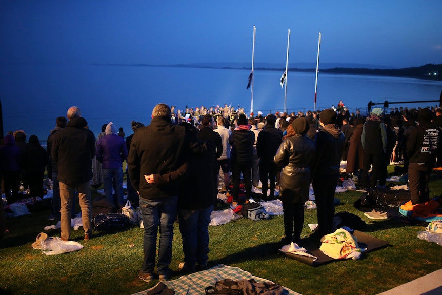 A crowd of people in warm clothes looks out over the early morning blue waters beyond three flags hung at half-mast