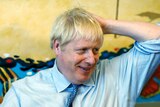 UK PM Boris Johnson wears a light blue shirt and holds his arm up to his head.