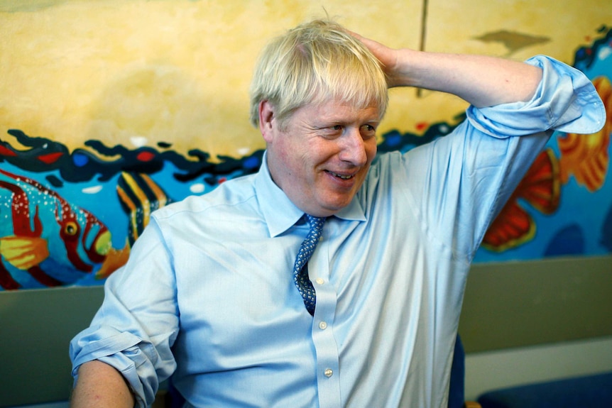UK PM Boris Johnson wears a light blue shirt and holds his arm up to his head.