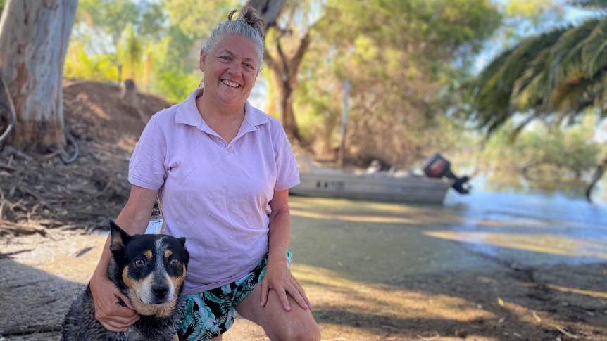 A white woman with grey hair and a pink shirt smiling, kneeling on the ground near her dog. River in background.