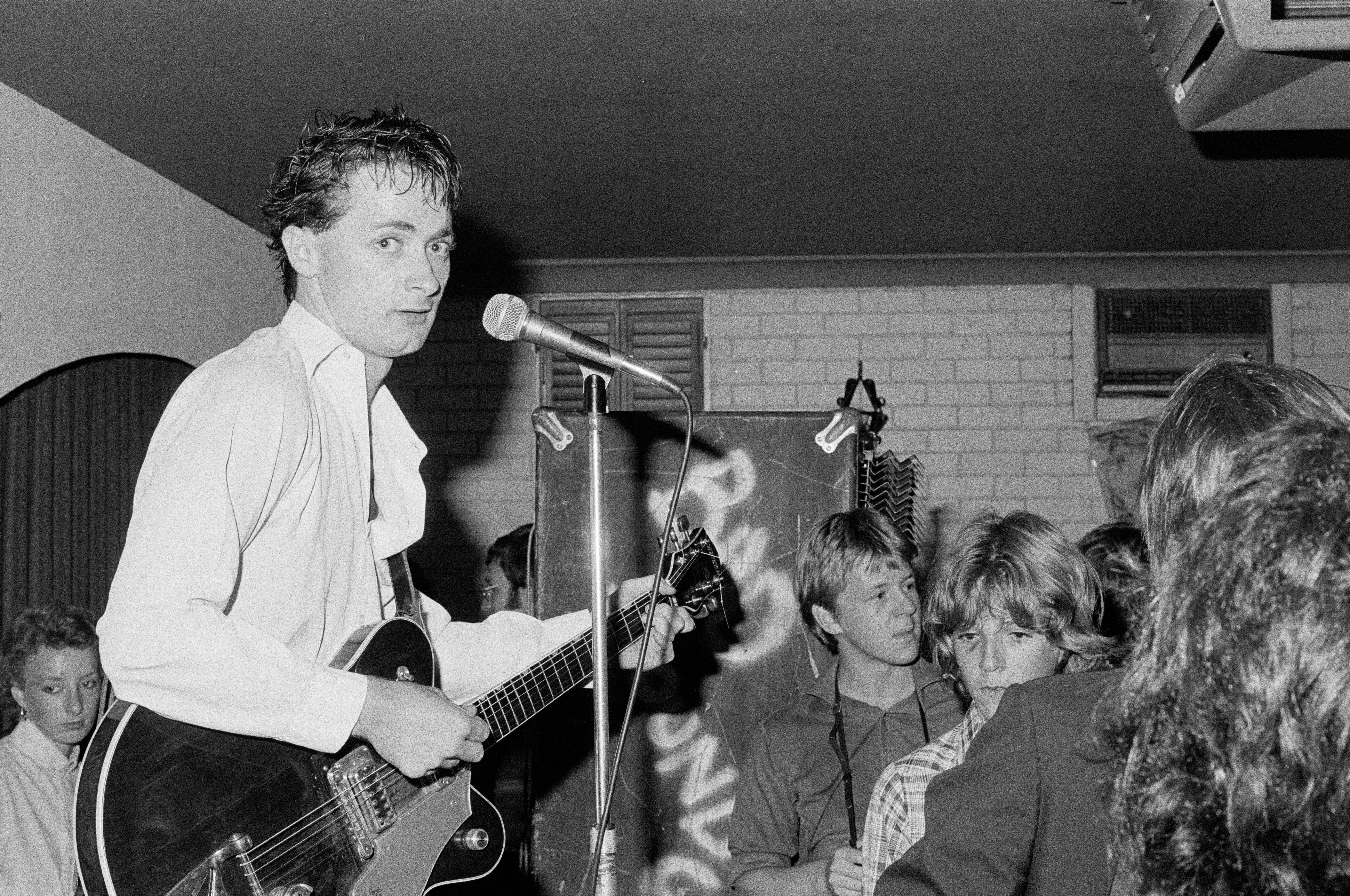 Black and white image of man in white shirt with guitar performing in front of a crowd