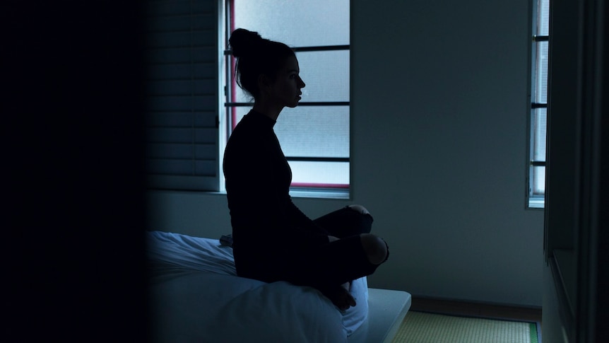 The silhouette of a woman sitting on end of bed. 