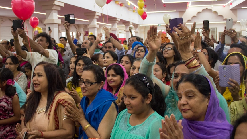 People raise hands, hold phones, smile and clap. Colourful dresses, balloons. High white ceiling and pillars.