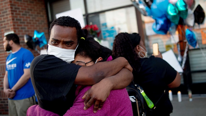 An African-American man and woman wearing masks hug each other surrounded by balloons and flowers.