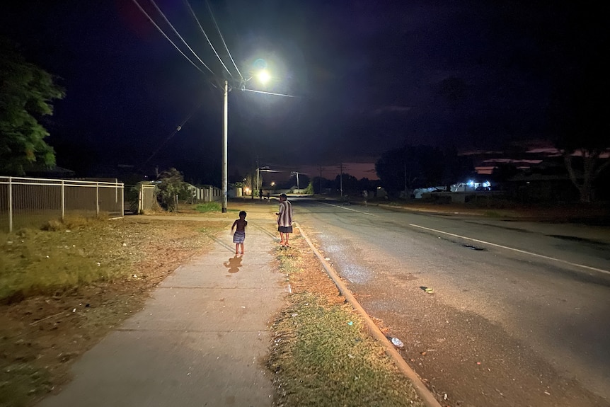 Two small children run down a street at night with a streetlight. 