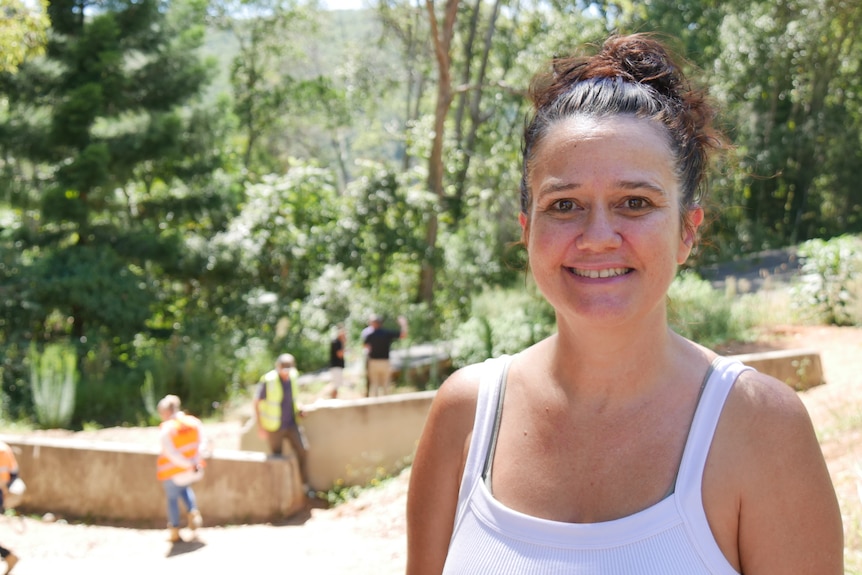 A woman wearing a ponytail is smiling as she stands in front of concrete barriers with workmen in the background