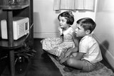 A black and white photo of a young boy and girl sitting on the floor looking at a radio. They are wearing clothes from 1940s.