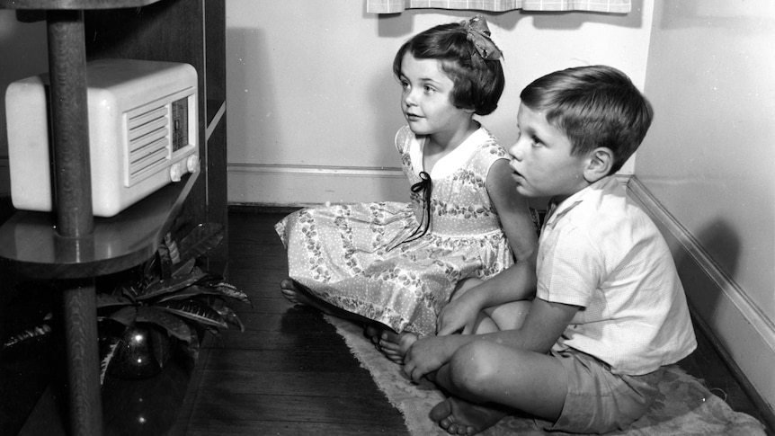 A black and white photo of a young boy and girl sitting on the floor looking at a radio. They are wearing clothes from 1940s.