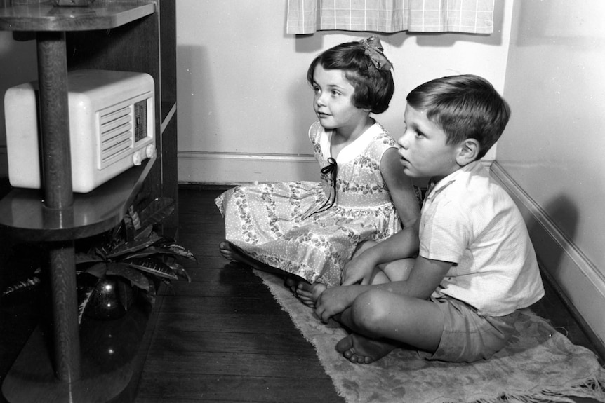 A black and white photo of a young boy and girl sitting on the floor looking at a radio. They are wearing clothes from 1940s.