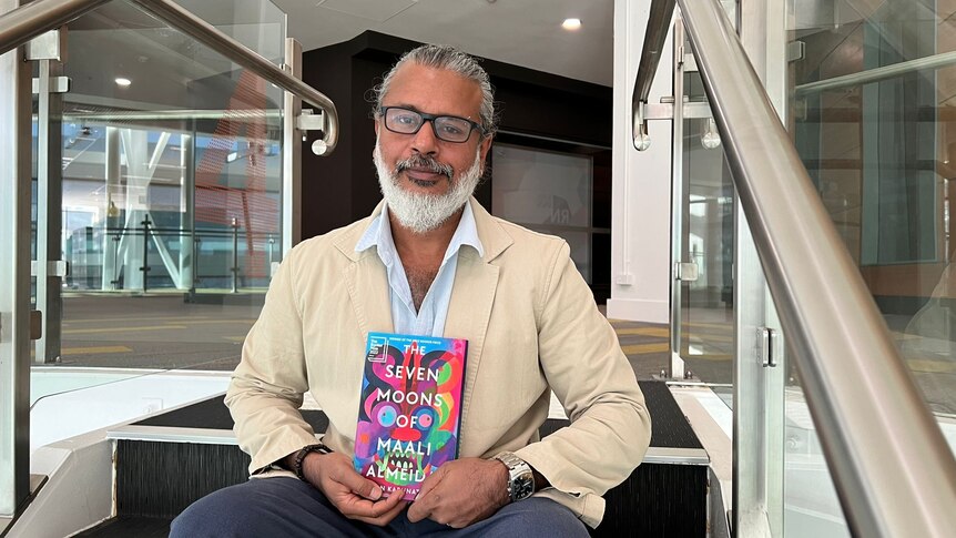 Shehan Karunatilaka author and winner of the manbooker prize sits down on stairs holiding book 'the seven moons of maali almeida