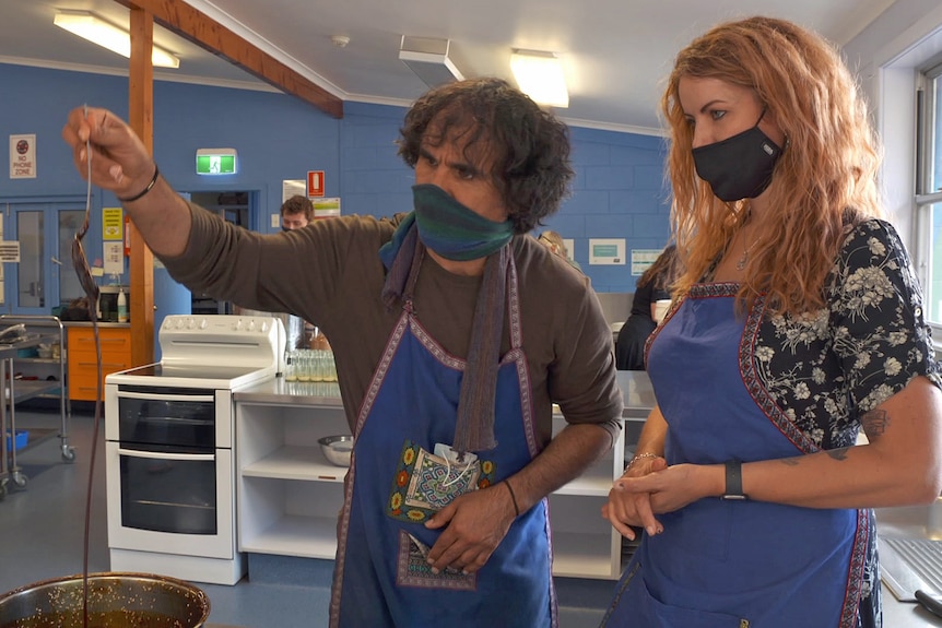 A man holds a spoon over a cooking pot. A woman stands next to him. Both wear face masks.