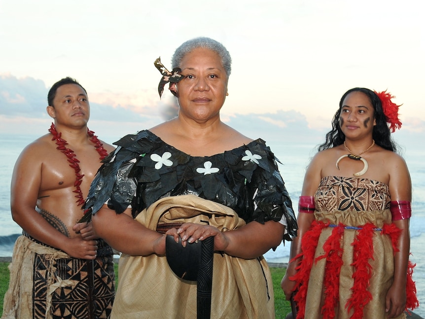 Fiame Naomi Mata'afa stands in front of a man and woman, all in traditional Samoan garments, on a beach at dusk.