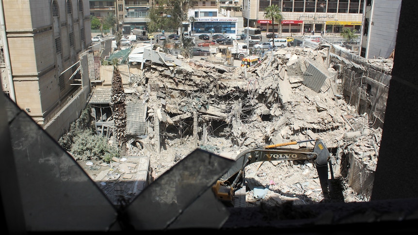 The rubble of a building sits in between two other standing buildings as an excavator begins to clear the rubble