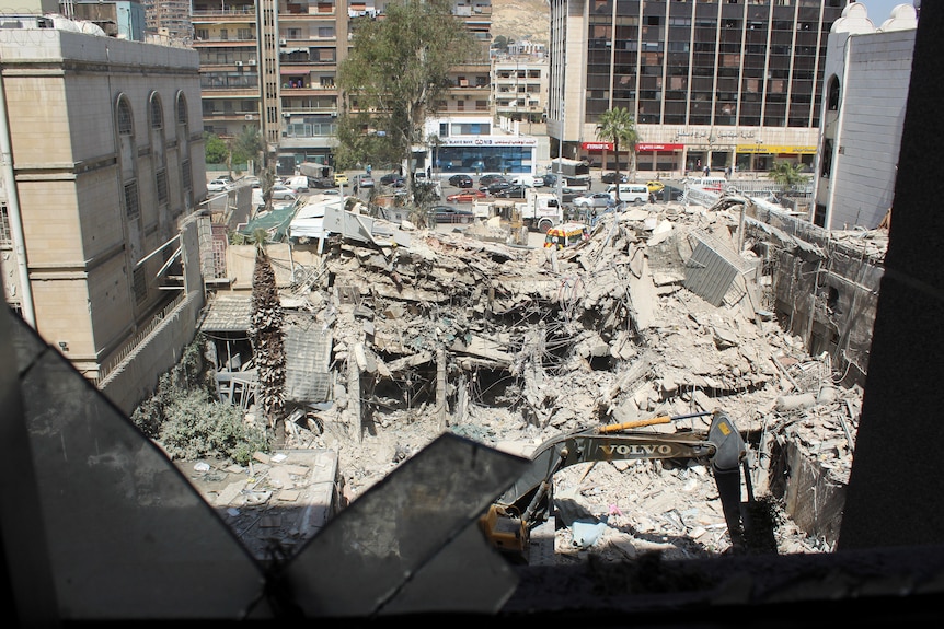 The rubble of a building sits in between two other standing buildings as an excavator begins to clear the rubble