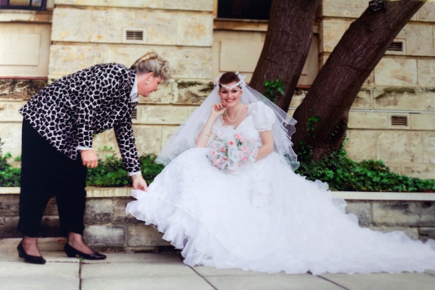 A woman in an animal print jacket bends to adjust the ruffled dress of a smiling bride seated on steps.