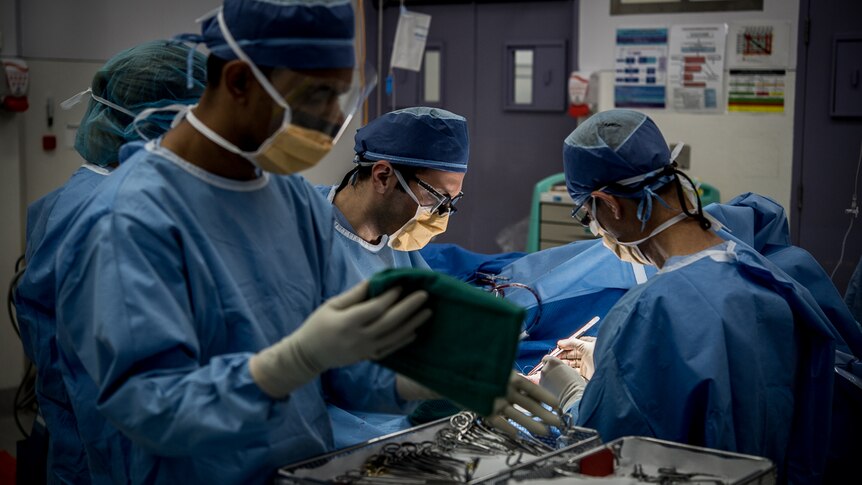 Four people dressed in surgical scrubs stand around a patient in an operating theatre.