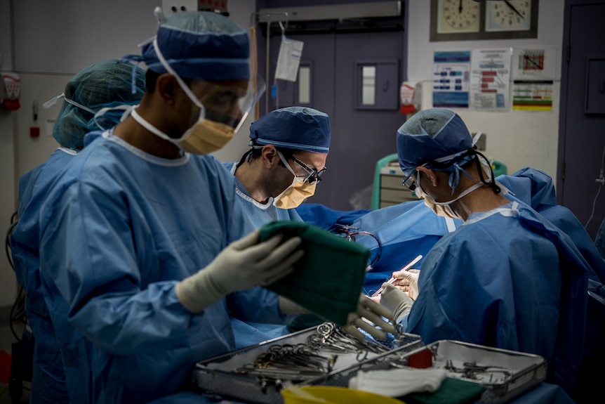 Four people dressed in surgical scrubs stand around a patient in an operating theatre.