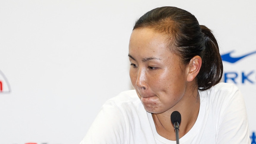 Peng Shuai looks to the side while sitting at a desk for a press conference with a microphone in front of her.