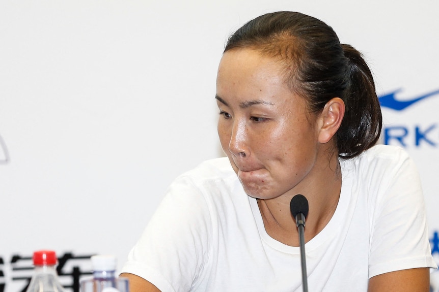 Peng Shuai looks to the side while sitting at a desk for a press conference with a microphone in front of her.