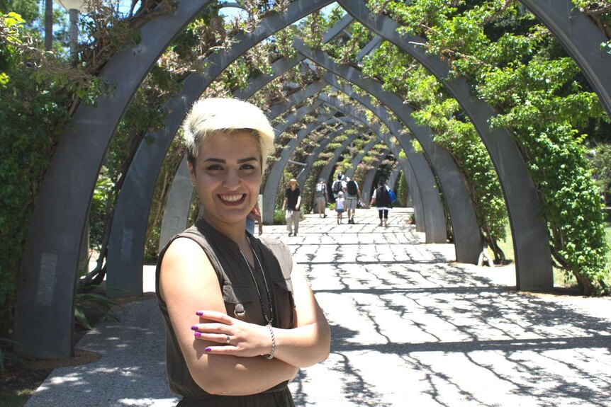 Mojgan stands under a pathway lined with trees with arms folded.
