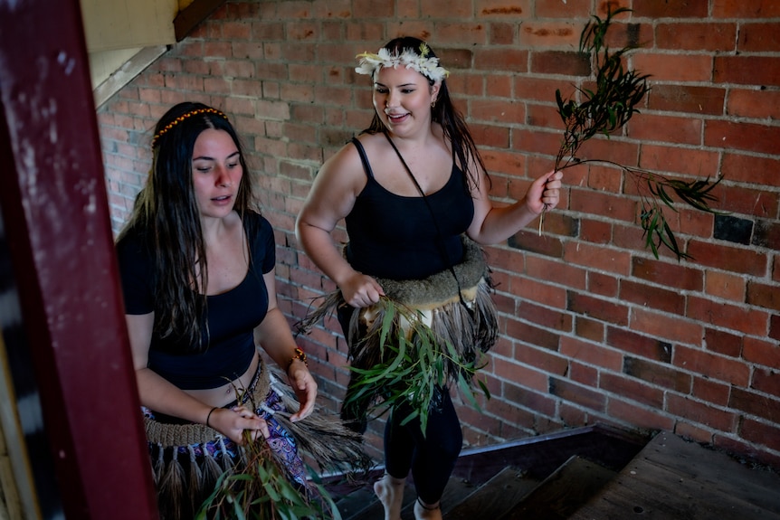 Two girls wearing feathered skirts and holding branches walk up a staircase smiling.