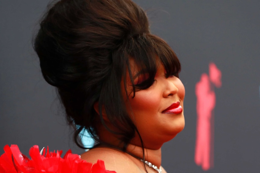 Lizzo smiles on the red carpet, wearing a red dress with feathers