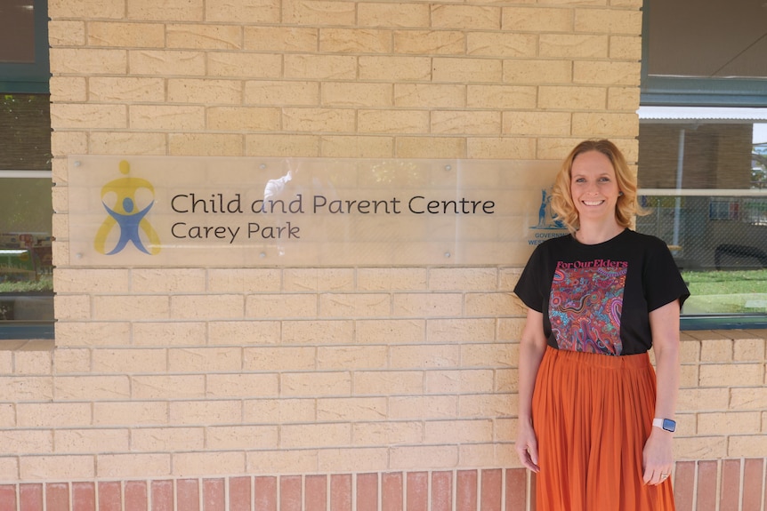 A woman with blonde hair, a black t-shirt and orange skirt stands next to the carey park child and parent centre sign.