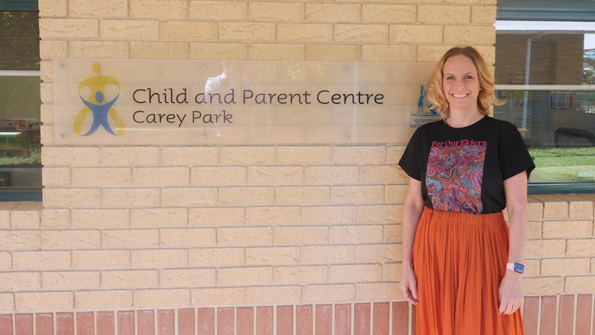 A woman with blonde hair, a black t-shirt and orange skirt stands next to the carey park child and parent centre sign.