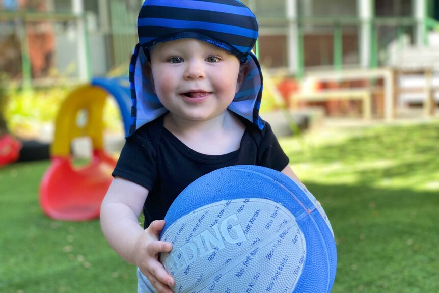 A toddler in a blue striped hat holds a basketball.