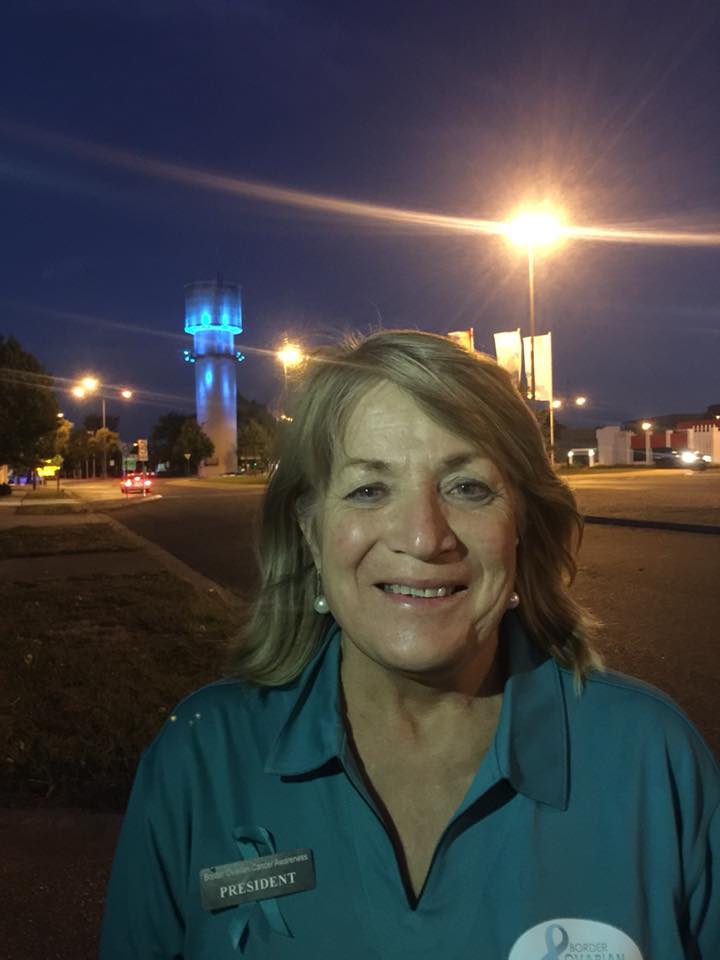 A blonde woman in a teal polo shirt stands at night in front of a lit up water tower in Wodonga. 