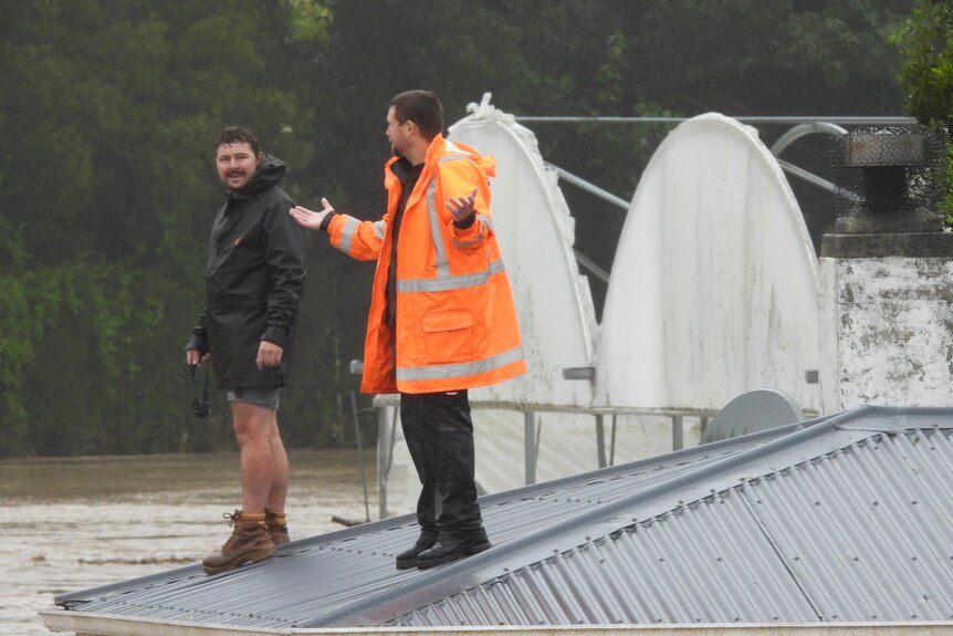 Two men standing on top of a roof with floodwaters around them. One is wearing hi-vis.