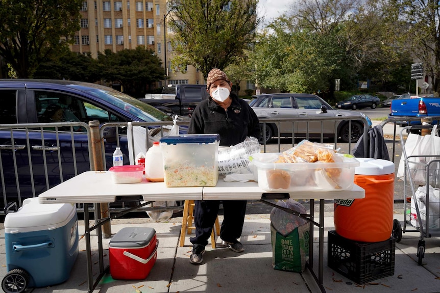 A man wearing a beanie and a white mask with a black jacket stands at a table covered in food containers