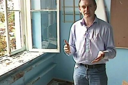 A man stands in a decrepit room gesturing to the camera.