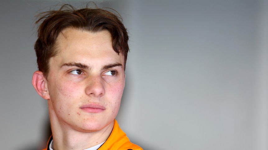 An F1 driver in an orange race suit, in the garage, looking over his left shoulder