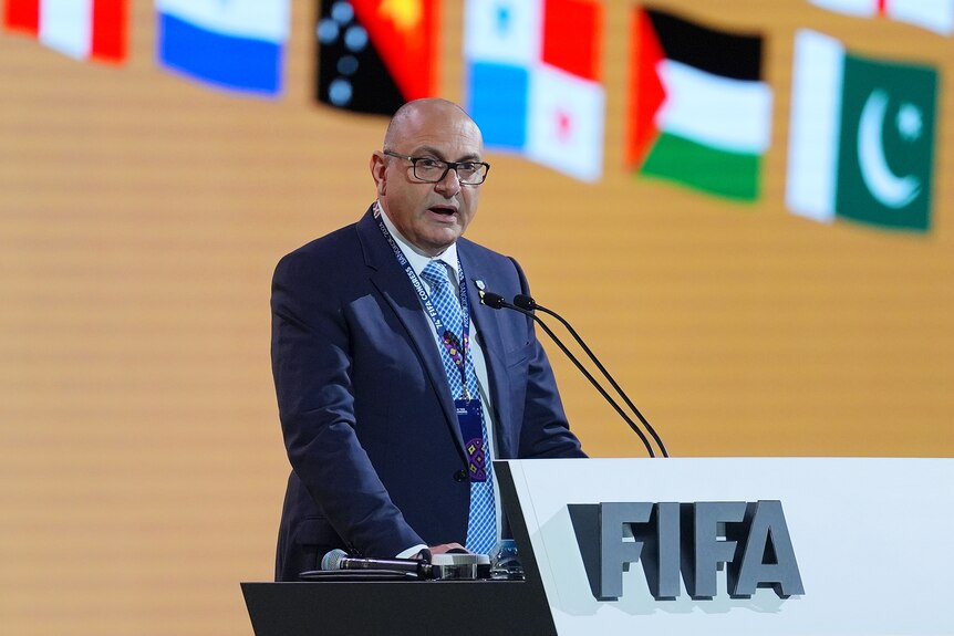 A bald man wearing glasses and a blue suit speaks at a lectern with national flags in the background
