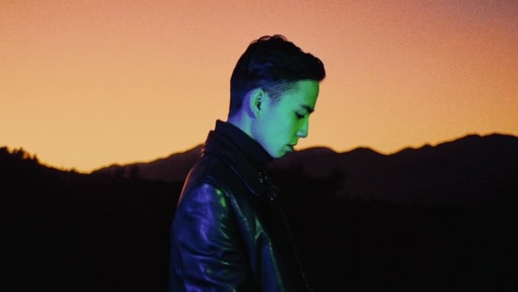 Manila faces right and is looking down. He wears a black leather jacket & a green light shines on his face. Sunset background.