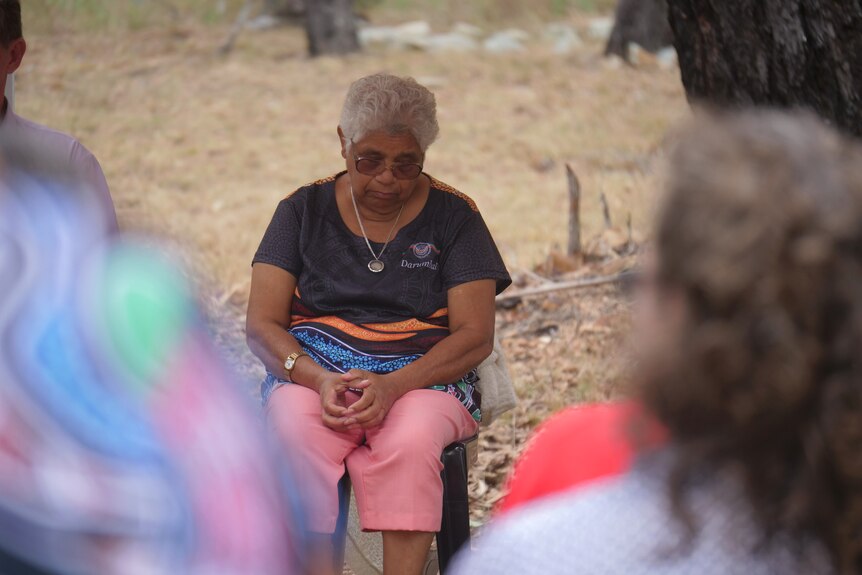 An Indigenous woman sitting in a chair, looking down with the hands sitting in her lap.