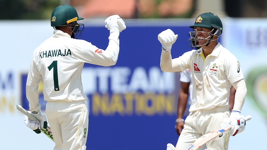 Dos jugadores de cricket de prueba australianos sonríen y se mueven para golpear los guantes en celebración después de una victoria.