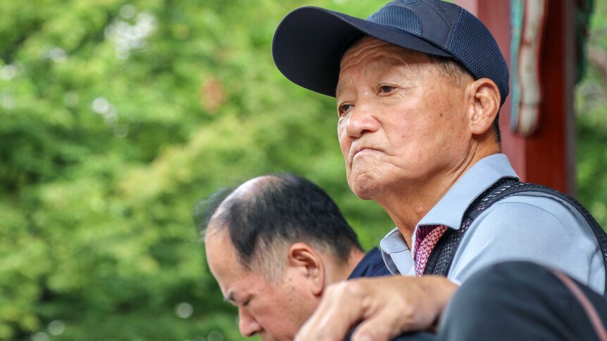 A elderly man in a baseball camps looks off frame against an unfocused leafy background